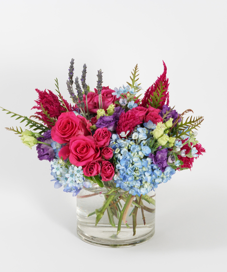 floral design of blue hydrangea, hot pink roses, and plum celosia arranged in a clear glass vase