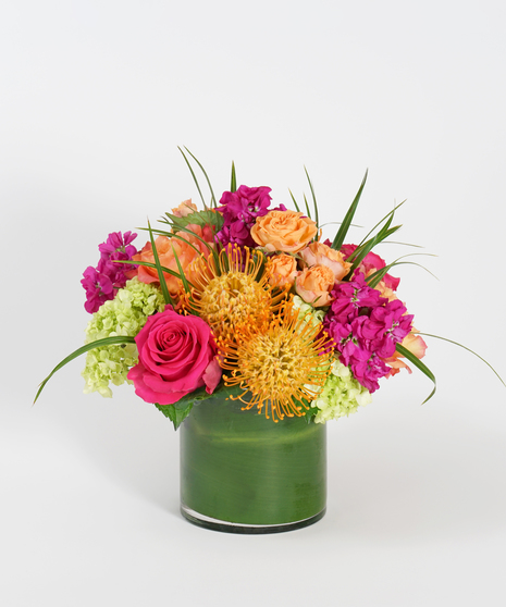 hot pink roses, orange protea, and fuchsia stock accented with greens in a leaf-lined glass vase