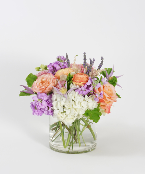 summer floral design of peach ranunculus, lavender clematis, and white hydrangea arranged in a clear glass vase