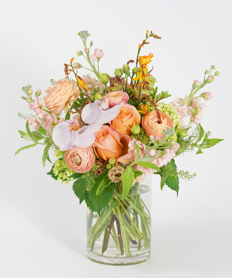 floral design of coral roses, peach ranunculus, and phalaenopsis orchids accented with orange crocosmia and arranged in a clear glass vase