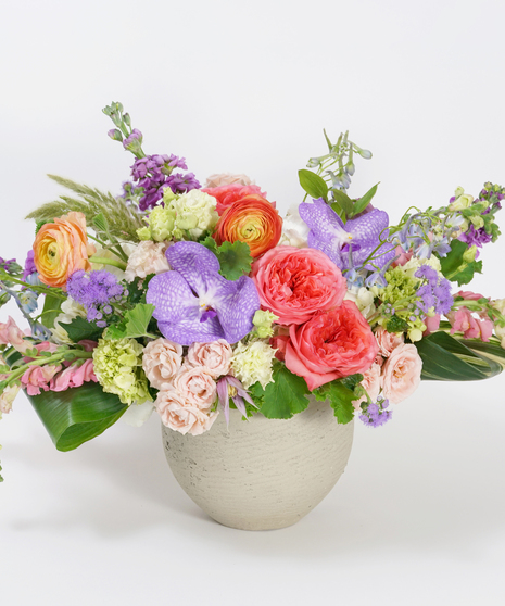 lush collection of lavender orchids, pink roses, and peach ranunculus arranged in a grey stone bowl
