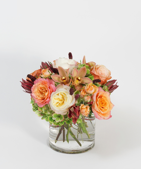 fall floral design of orange roses, coral spray roses, and bronze orchids arranged in a clear glass vase