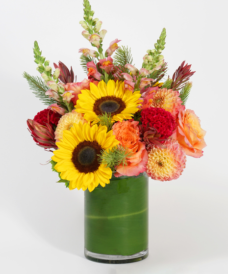 colorful floral design featuring sunflowers, orange roses, and peach dahlias accented with seasonal textures