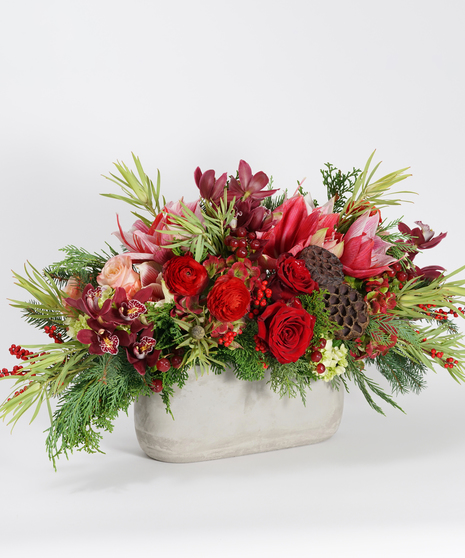 unique Christmas centerpiece of two-toned amaryllis, red ranunculus, and burgundy orchids arranged in a grey stone container and accented with seasonal greens