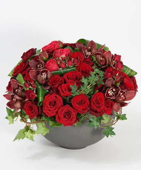 lush mound of red roses accented with burgundy orchids and ivy arranged in a large grey stone bowl