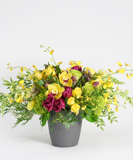 yellow and purple floral design featuring orchids, tulips, and roses arranged in a dark stone container