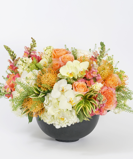 luxury floral design of yellow orchids, orange roses, snapdragon, and protea accented with variegated dracaena and designed in a grey stone bowl