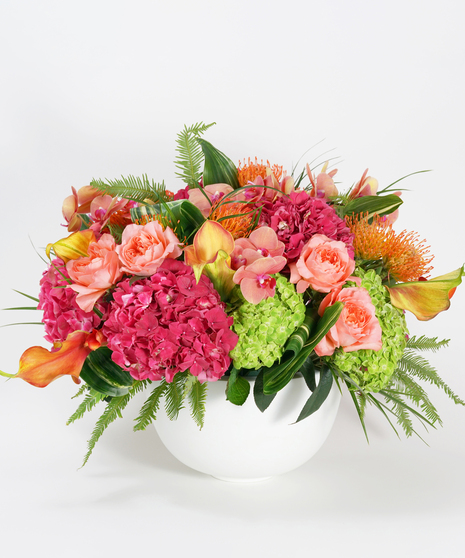 luxury floral design of coral orchids, mango calla lilies, snapdragon, and protea accented with ferns and designed in a white stone bowl