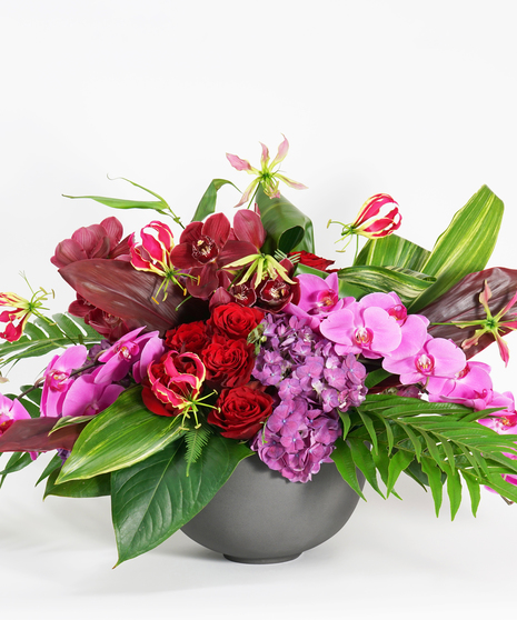 luxury floral design of pink orchids, purple orchids, red roses, and purple hydrangea arranged in a grey stone bowl