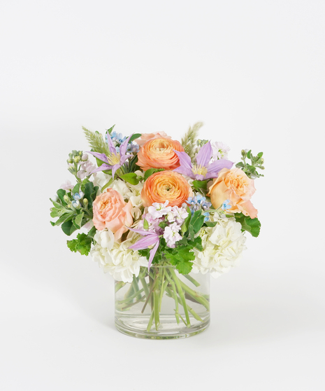 summer floral design of peach ranunculus, lavender clematis, and white hydrangea arranged in a clear glass vase