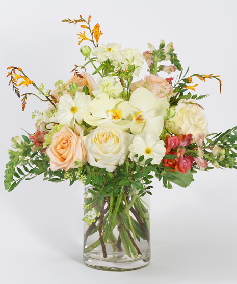 gardeny summer floral design of soft yellow orchids, peach roses, and orange snapdragon arranged in a clear glass vase and accented with seasonal greens