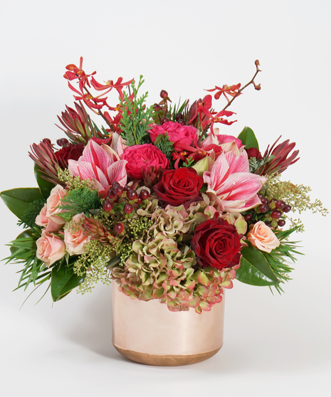 unique holiday floral design of two-toned amaryllis, peach roses and burgundy orchids arranged in a copper container and accented with winter greens