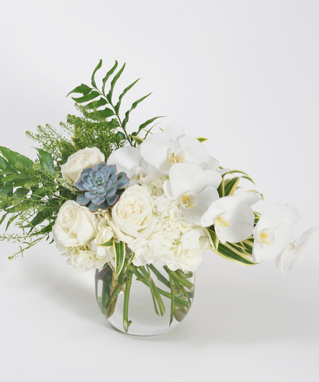 asymmetrical white floral design of white orchids, roses and hydrangea arranged in a glass bowl and accented with textural greens